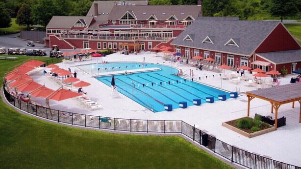 Una amplia piscina con varias pistas de natación, rodeada por una zona cubierta con asientos y sombrillas, y un edificio con techo rojo y detalles blancos al fondo.