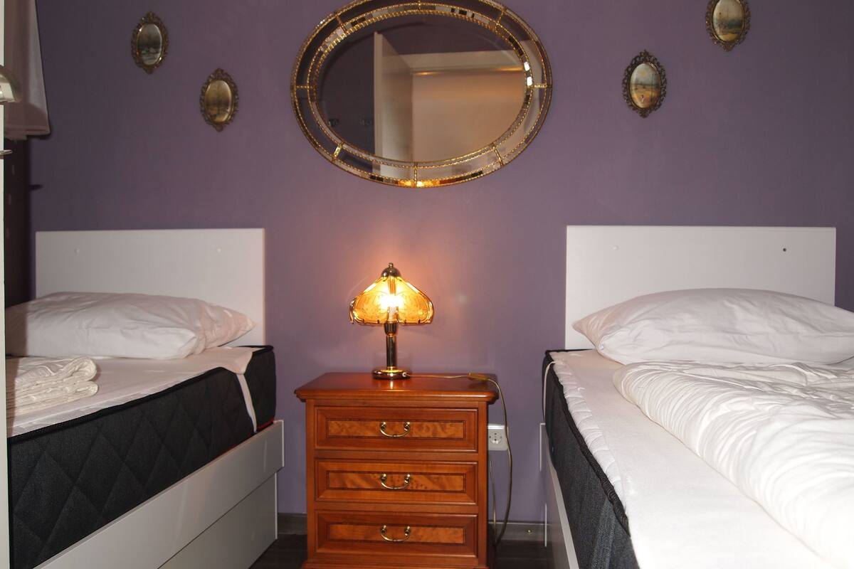 A hotel room with two beds, a wooden nightstand with a lamp, and a round mirror on the wall.