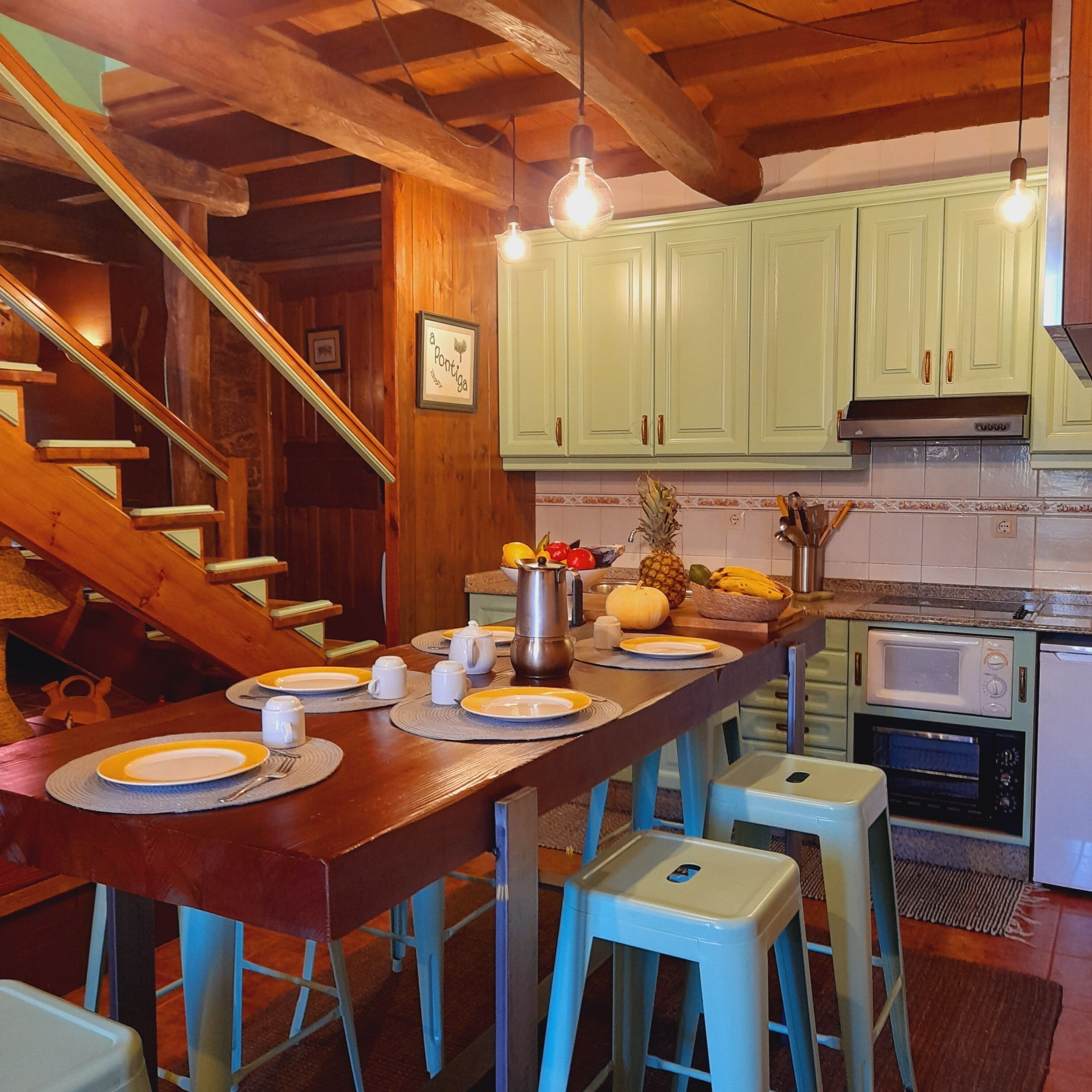 Una cocina con vigas de madera, armarios verdes, una mesa de comedor con platos y tazas, y un microondas.