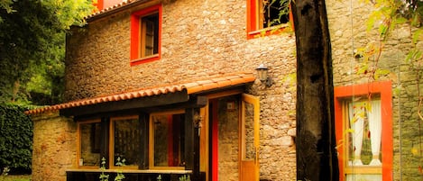 A stone house with a tiled roof, wooden shutters, and a small porch with a bench.