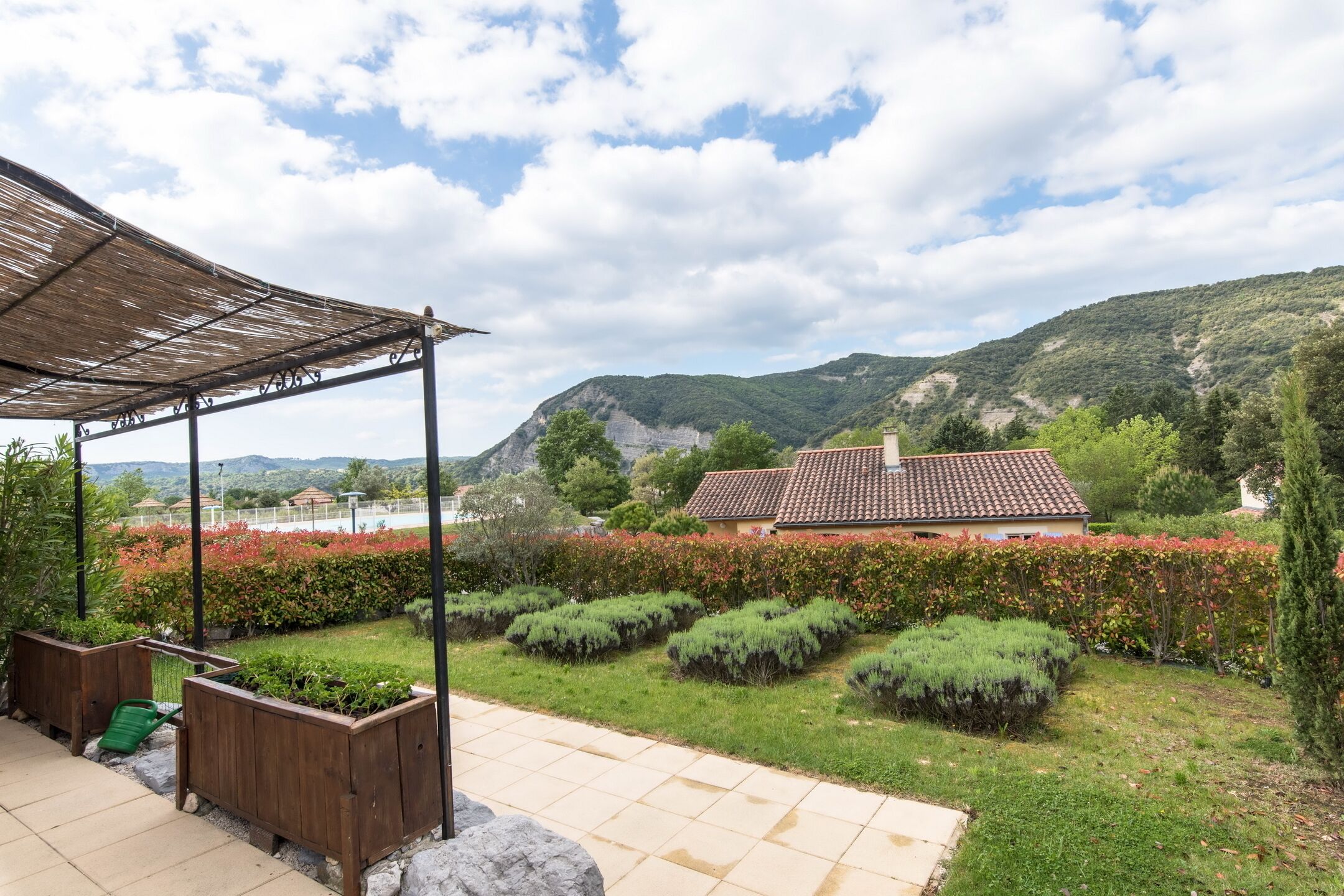 Un jardín con un pérgola, setos recortados y una casa con tejado de tejas al fondo.