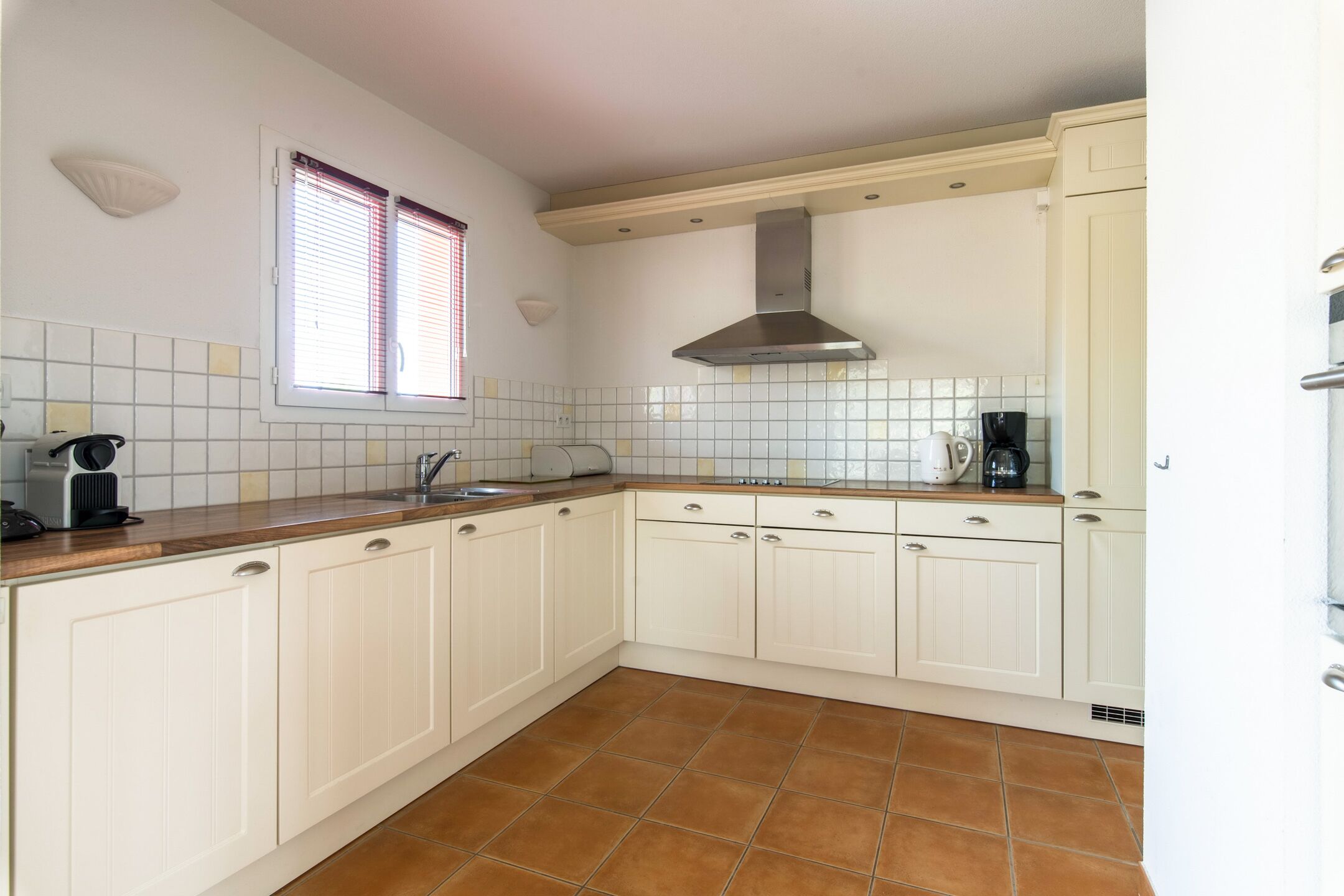 Una cocina con armarios blancos, una campana extractora de acero inoxidable, una ventana con persianas y un salpicadero de azulejos.