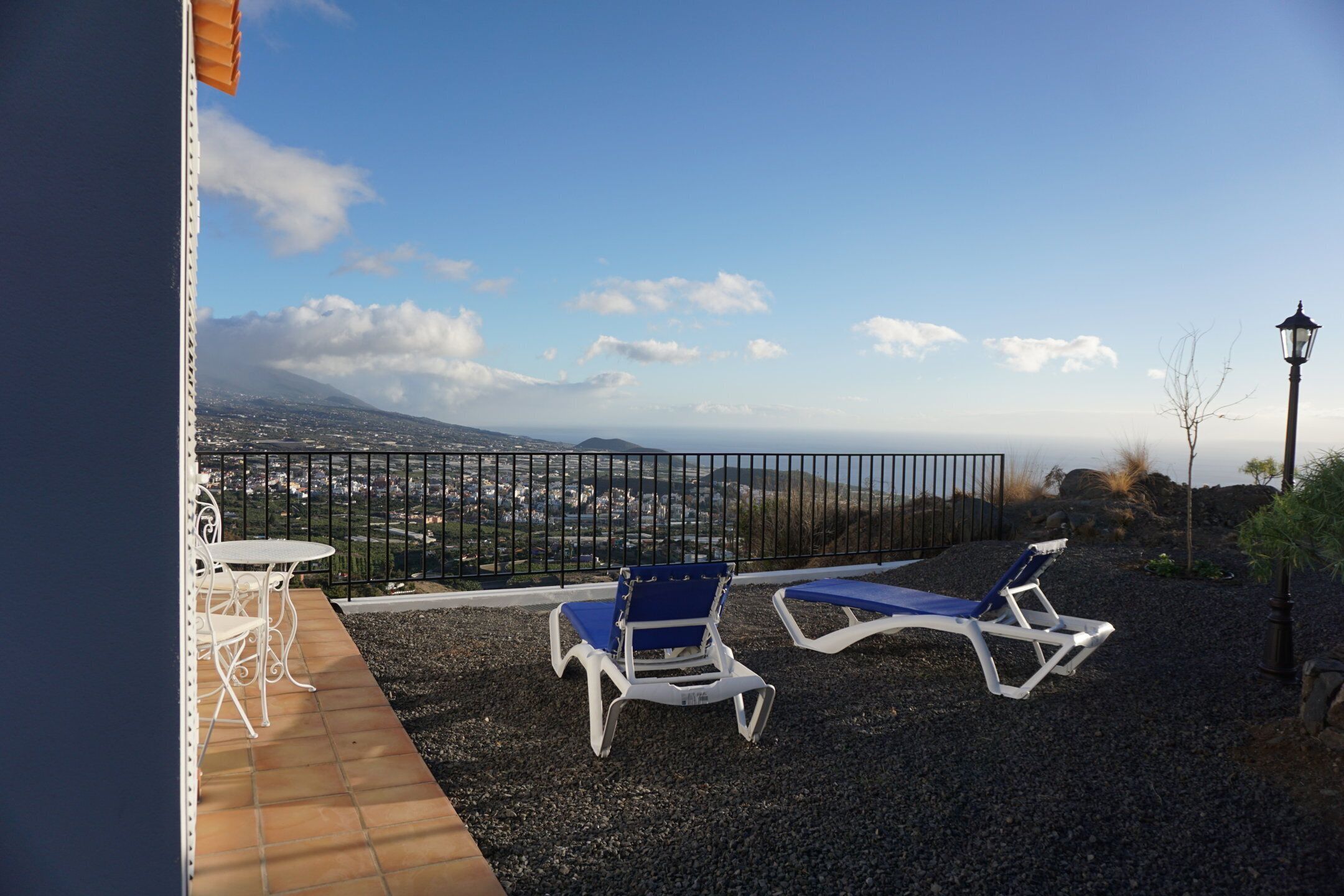 Una terraza con dos sillones azules y una mesa blanca, con vistas a un paisaje pintoresco que incluye una montaña y una ciudad a lo lejos.