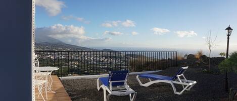 Una terraza con dos sillones azules y una mesa blanca, con vistas a un paisaje pintoresco que incluye una montaña y una ciudad a lo lejos.