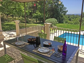 Un área de comedor al aire libre con una mesa dispuesta para dos, con vista a una piscina y un jardín cuidado.