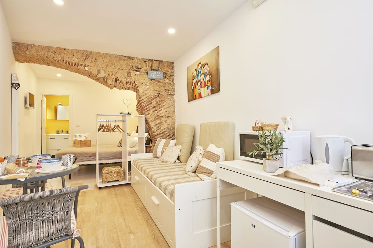 A modern, well-lit room with a bed, a desk, and a chair. There is a brick wall and a painting on the wall.