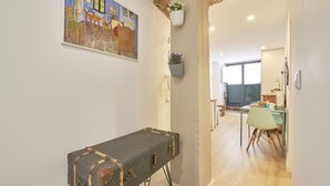 Una habitación con una mesa cubierta por una maleta antigua, un cuadro en la pared y vista a otra estancia con comedor.