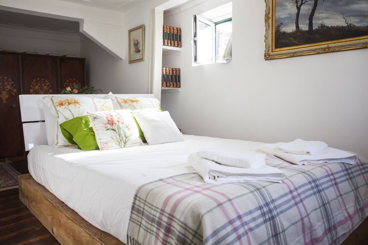 A neatly made bed with a plaid blanket, a window with a view of trees, and a bookshelf with books.