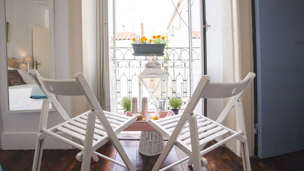 Una habitación con balcón, una silla blanca, una mesita con un adorno y vista a la calle.