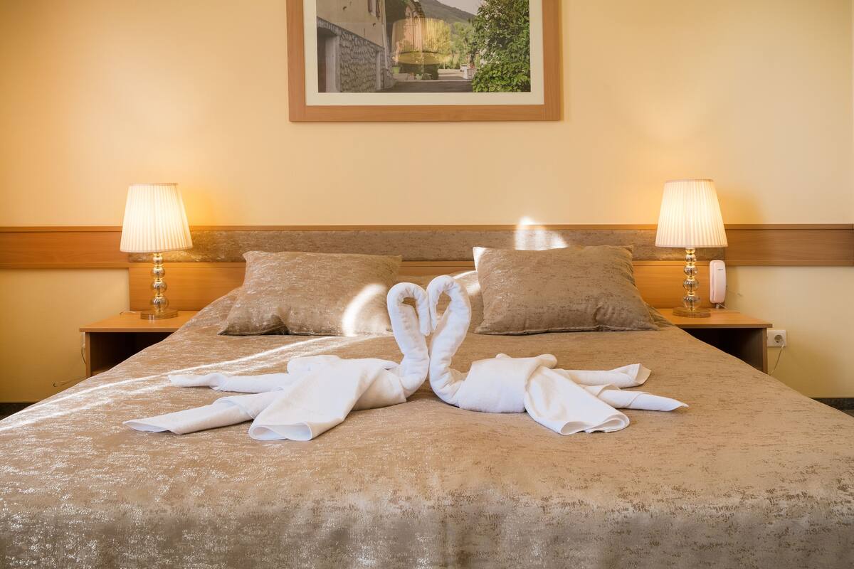 A neatly made bed with towels arranged in a swan shape, two bedside lamps, and a framed picture on the wall.