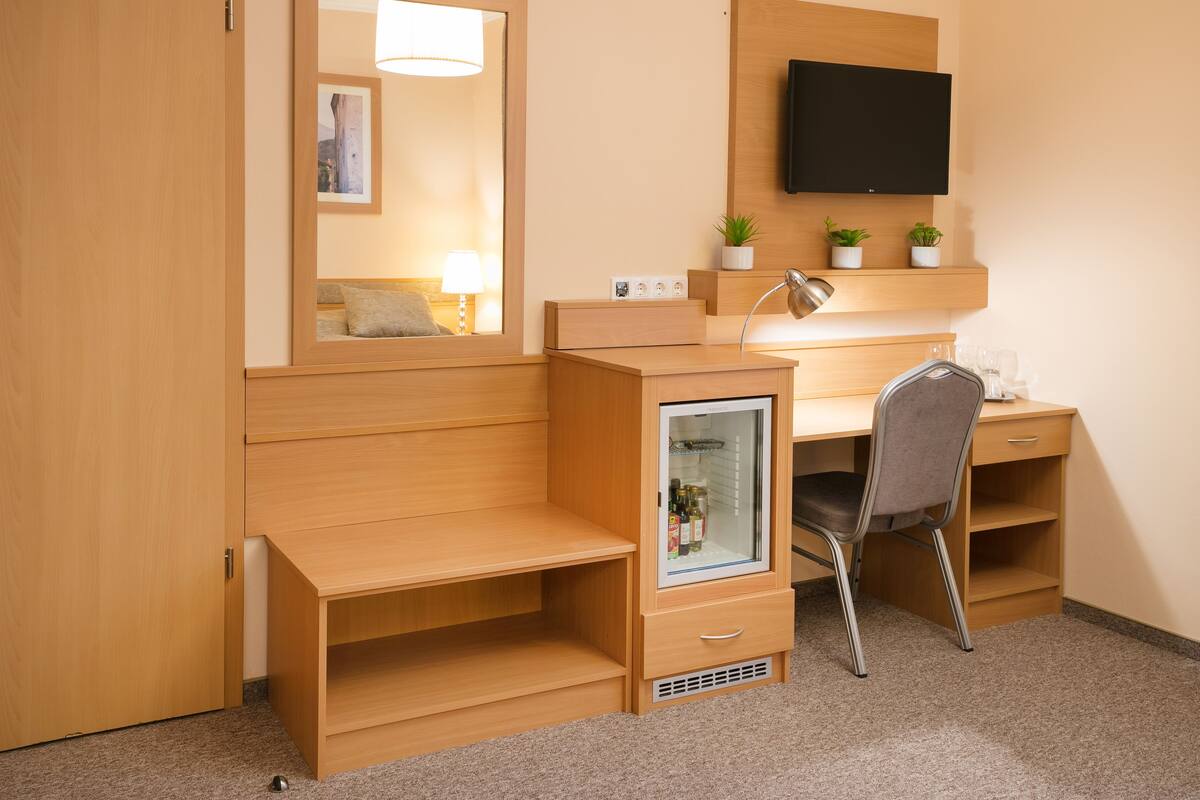 A compact hotel room with a wooden desk, a chair, a small fridge, a mirror, a flat-screen TV, and a bedside table.