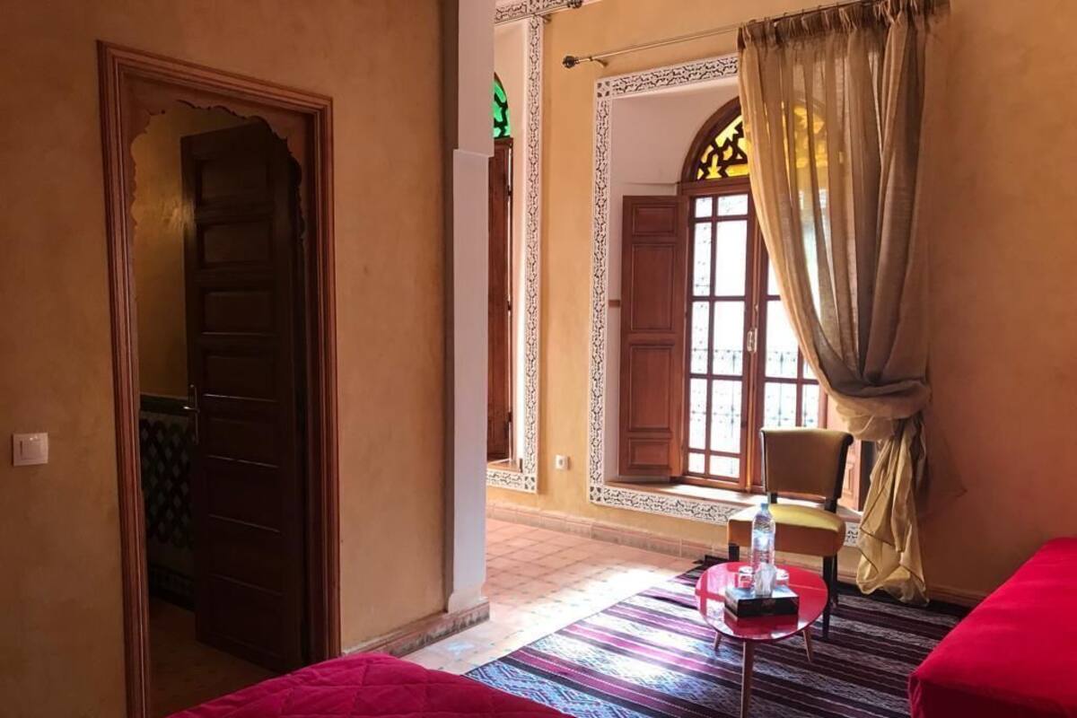 A hotel room with a bed, a small table with a bottle, a chair, and a window with curtains.