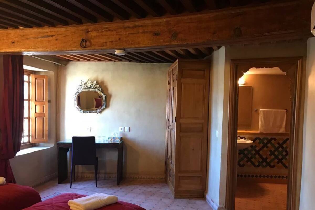 A room with a wooden ceiling, a bed with a red blanket, a desk with a chair, a mirror, and a bathroom with a patterned floor.