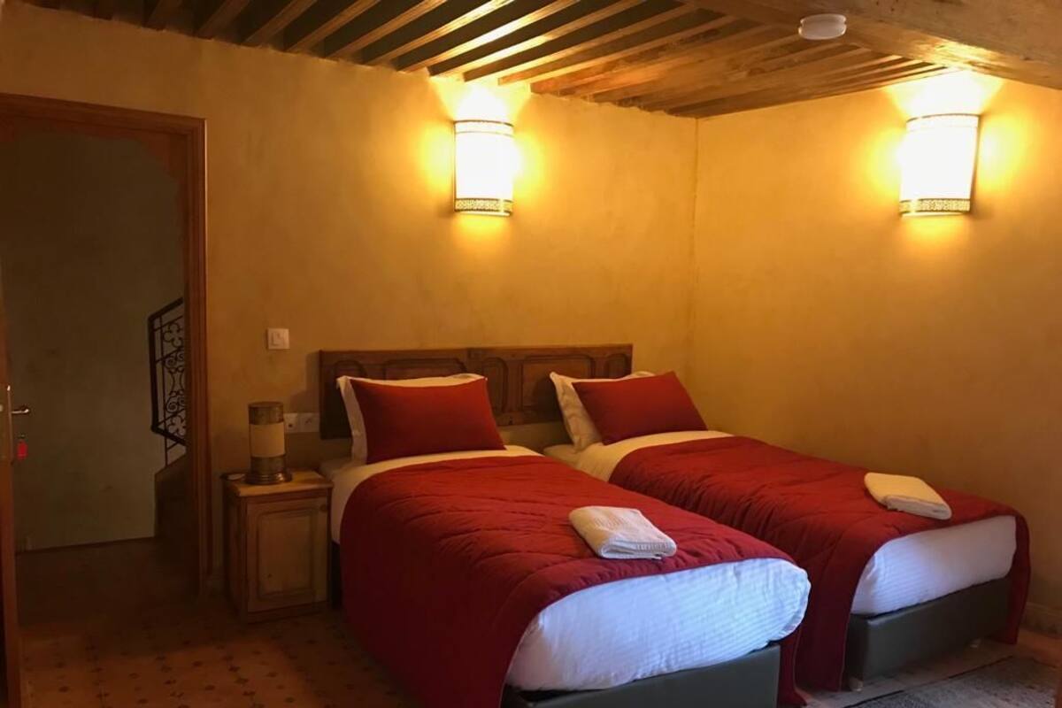A room with two beds, each with red bedding and white pillows, a small bedside table with a lamp, and two wall-mounted light fixtures.