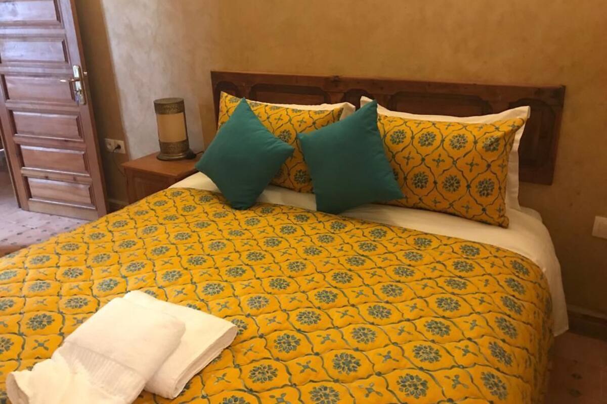 A bed with a yellow patterned bedspread, two teal pillows, and a white towel on the side.