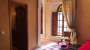 A hotel room with a red bed, a small table with a bottle, and a chair. There is a door leading to another room and a window with curtains. - Riad Jasmins & Spa (Fes)