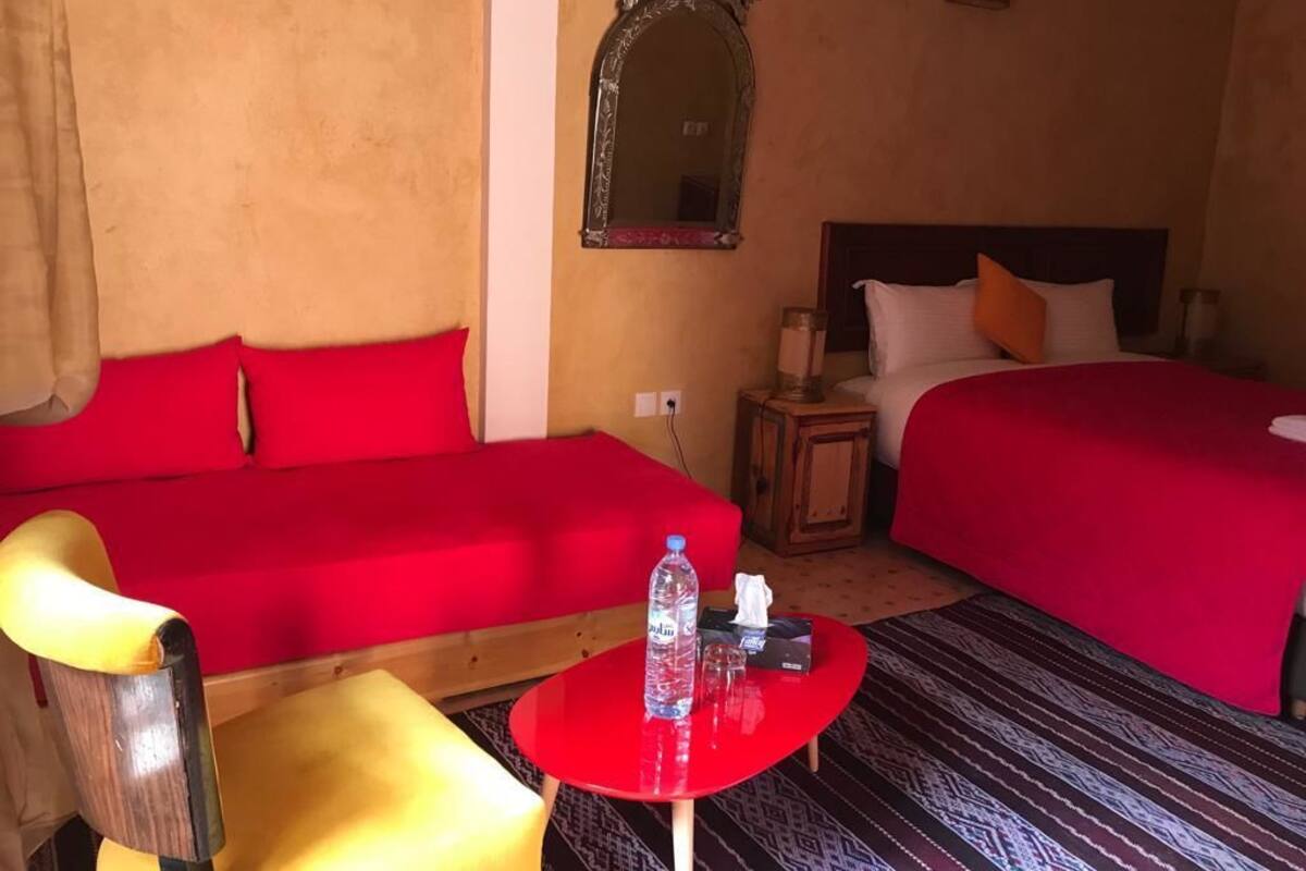 A hotel room with two red beds, a wooden chair, a small red table with a bottle and tissues, and a mirror on the wall.
