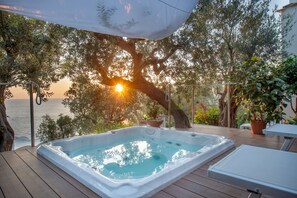 Uma banheira de hidromassagem em deck de madeira com vista para o mar e árvores ao pôr do sol.