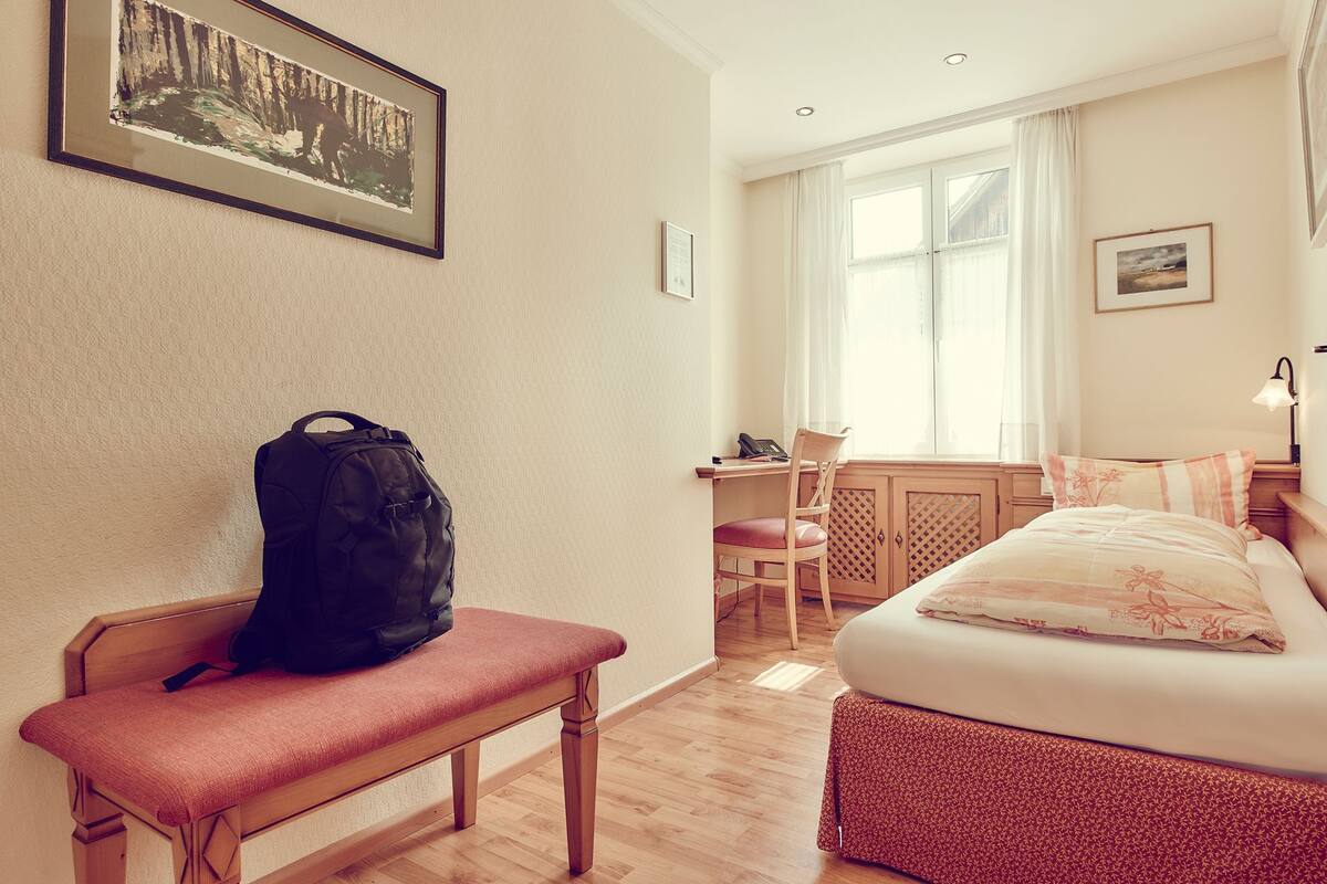 Ein Hotelzimmer mit einer hölzernen Bank, einem Schreibtisch mit Stuhl, einem Bett mit gemustertem Überwurf und einem Fenster mit Vorhängen.