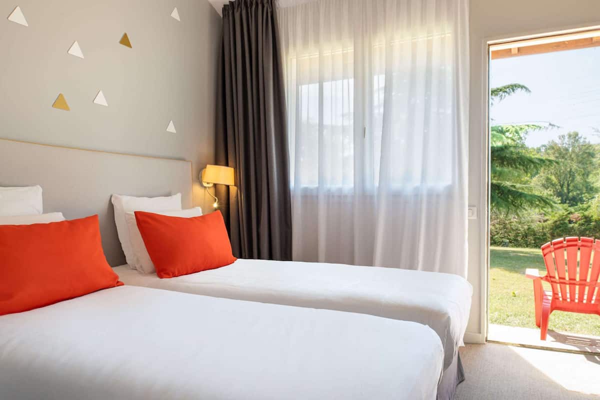Une chambre d’hôtel avec deux lits, une table de chevet avec une lampe, une fenêtre avec des rideaux légers et une vue sur la verdure.