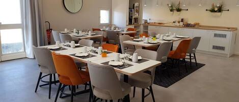 A dining area with multiple tables and chairs, a kitchen in the background, and a large window with curtains.