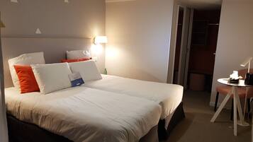 A hotel room with a large bed, a bedside table with a lamp, a small table with a coffee maker, and a chair.