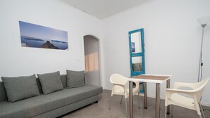 Una habitación con un sofá gris, dos sillas blancas, una mesita pequeña, un espejo con marco azul y un cuadro de un paisaje.