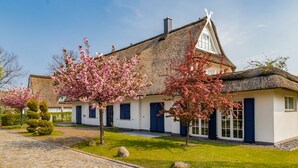 Een traditioneel huis met rieten dak, een witte gevel en blauwe luiken, omgeven door verzorgde tuinen en een keienpad.