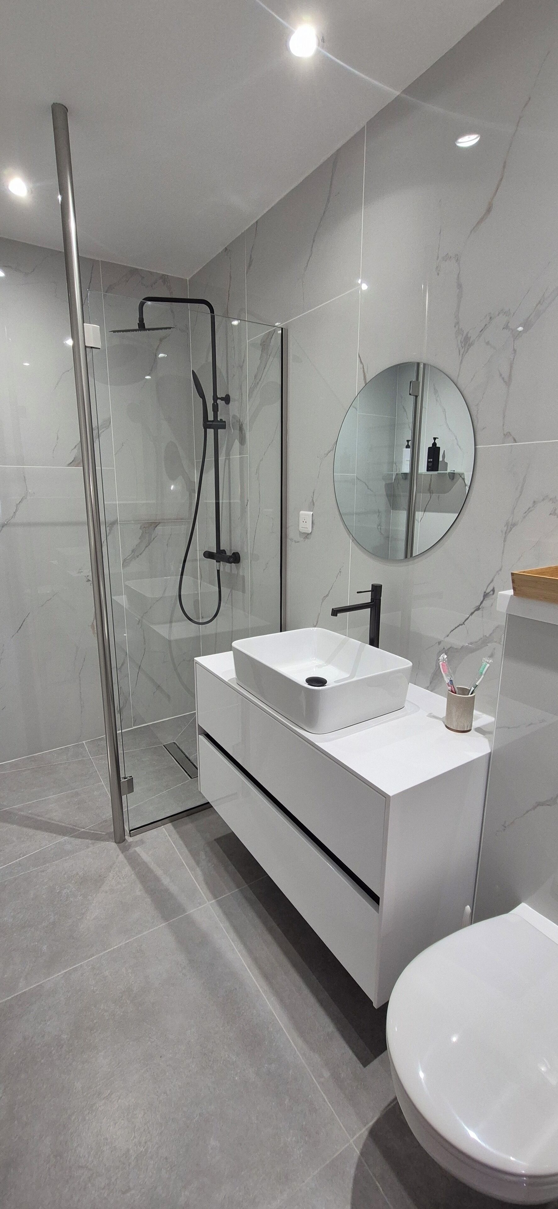 Un baño moderno con ducha envuelta en vidrio, tocador blanco con lavamanos y espejo redondo.