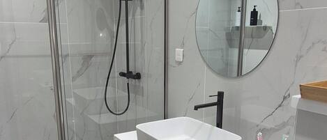 Un baño moderno con ducha envuelta en vidrio, tocador blanco con lavamanos y espejo redondo.