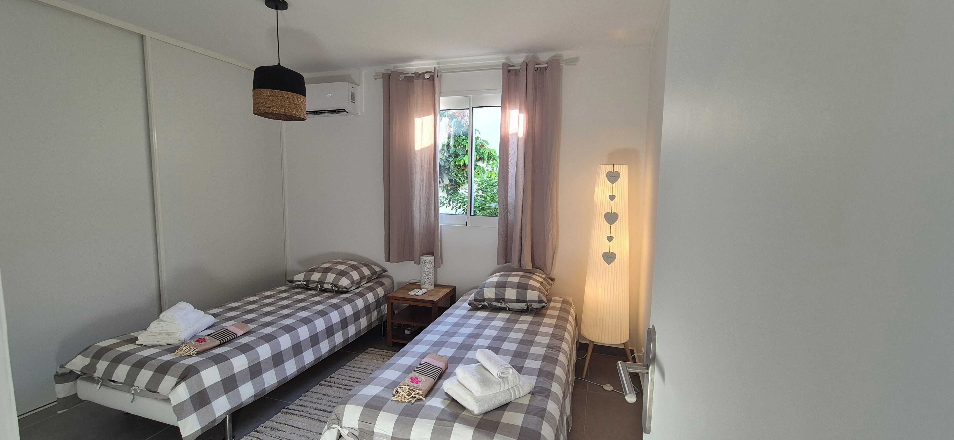 A small, clean room with two beds, a nightstand, a lamp, and a window with curtains.