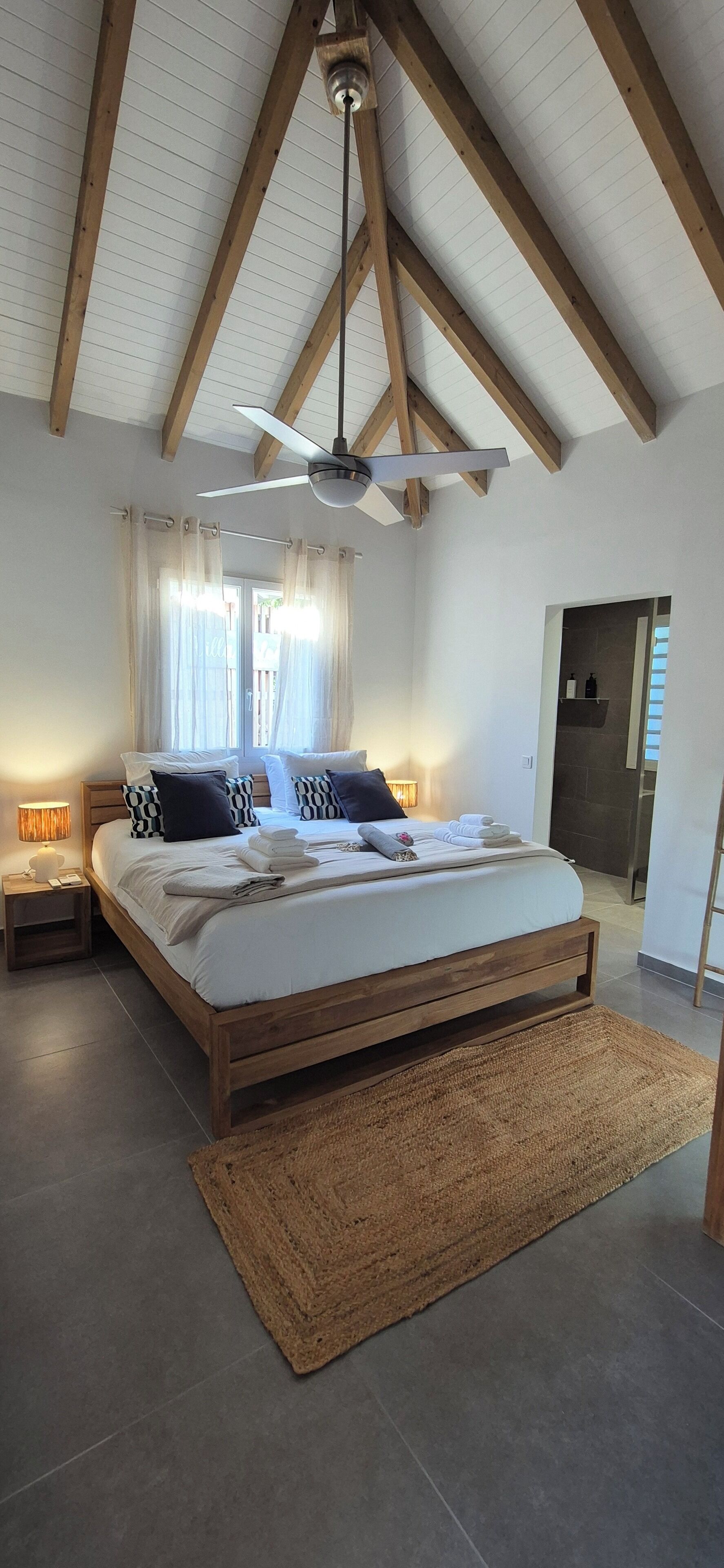 Een slaapkamer met een houten bed, een plafondventilator en een groot raam met gordijnen.