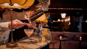 Un bar con mostrador de mármol, un cóctel en preparación y una vela encendida sobre una mesa pequeña al fondo.