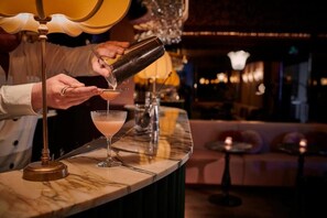 Un bar con mostrador de mármol, un cóctel en preparación y una vela encendida sobre una mesa pequeña al fondo.