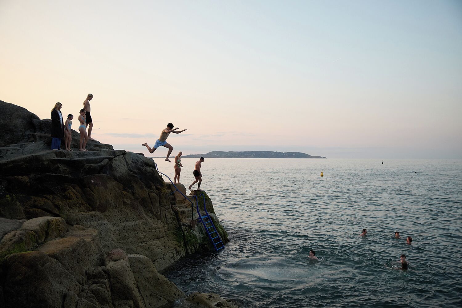 A rocky coastline with a ladder leading into the sea, a group of people on the rocks, and swimmers in the water.