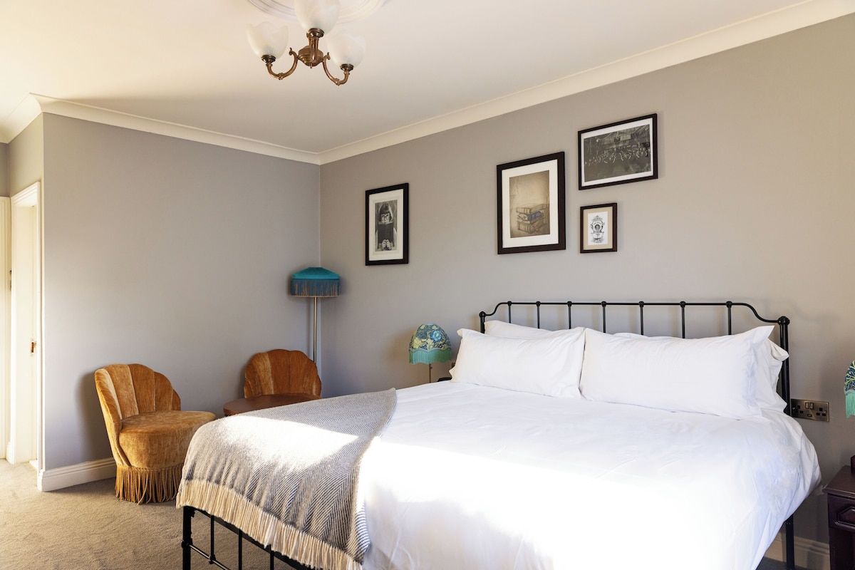 Un dormitorio con una cama, dos sillas, una lámpara y tres cuadros en la pared.