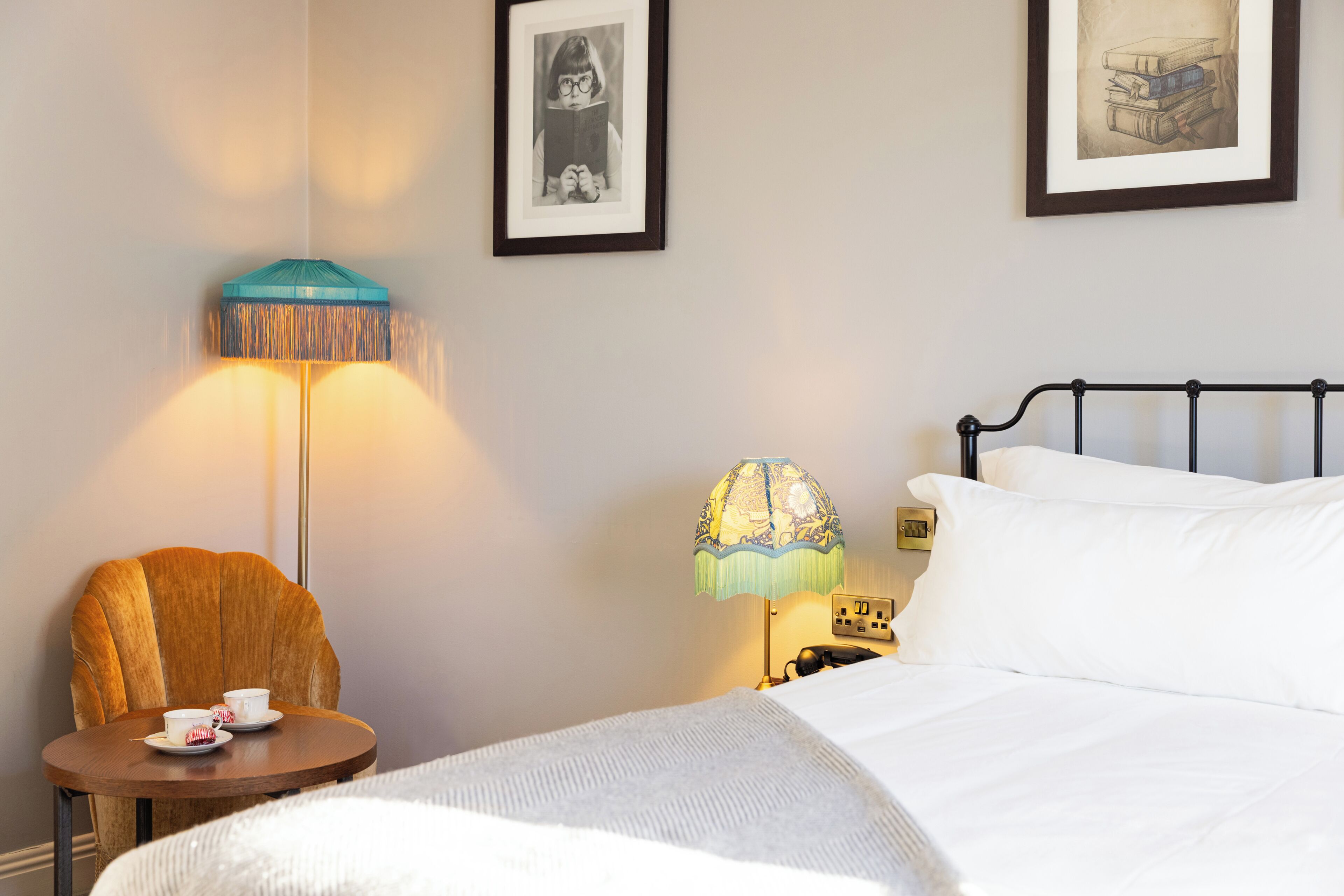 A hotel room with a bed, a chair, a table with a cup, two lamps, and two framed pictures on the wall.