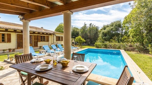 Uma área externa coberta com piscina, mesa de jantar posta com pratos e talheres, e cadeiras de descanso.