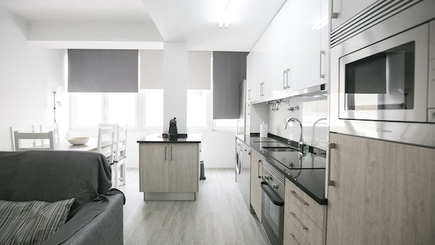 A modern kitchen with stainless steel appliances, a central island, and a grey sofa in a bright, open-plan living area.
