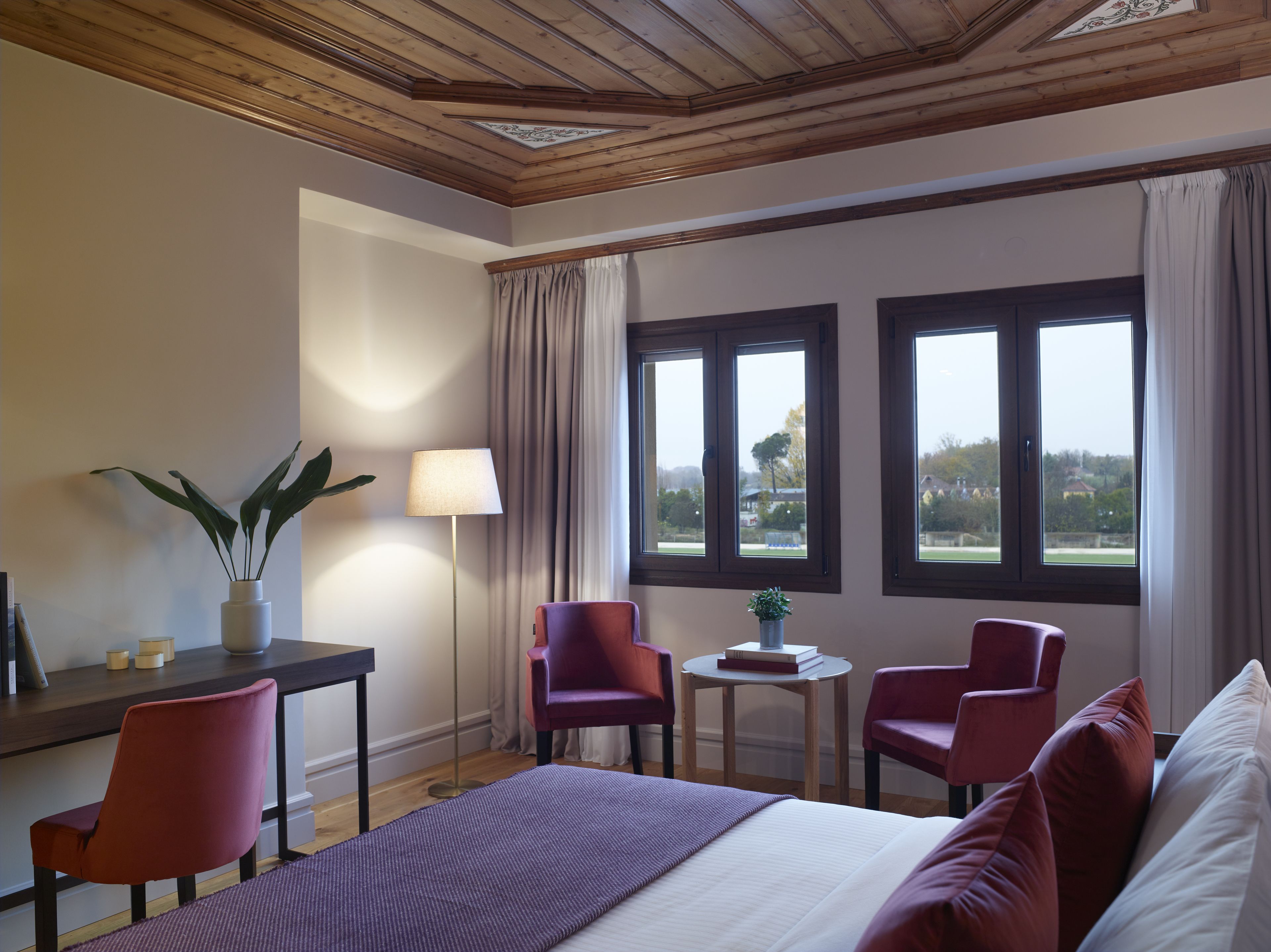 A hotel room with a bed, two chairs, a small table, a lamp, and a view of the outdoors through two windows.