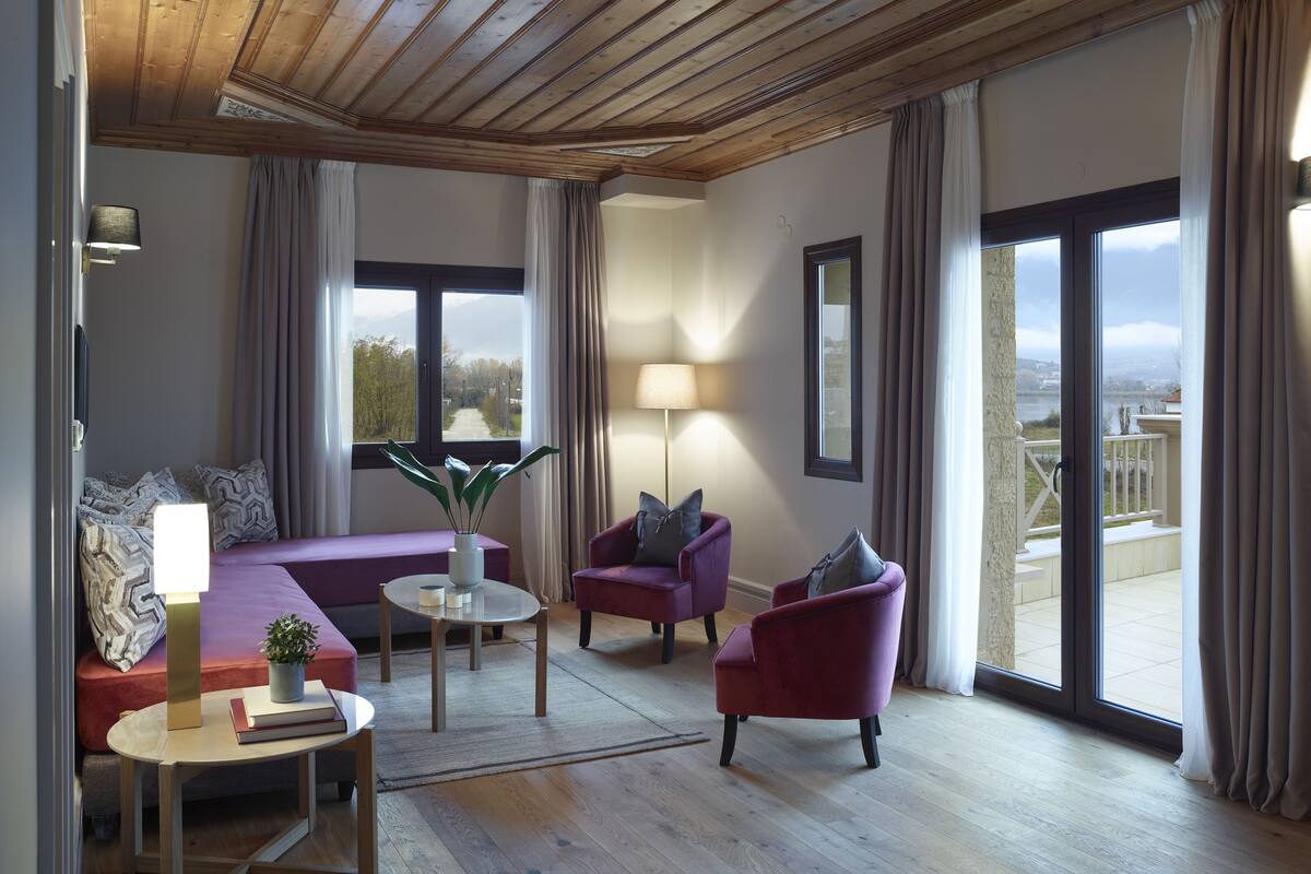A spacious living room with a wooden ceiling, large windows with curtains, a sofa, a chair, a small table with a lamp, and a view of a river and mountains.