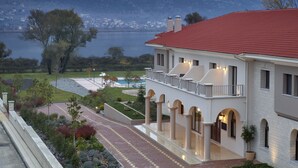 A large, two-story building with a red roof, a swimming pool, and a view of a lake. - The Lake Hotel (Ioannina)