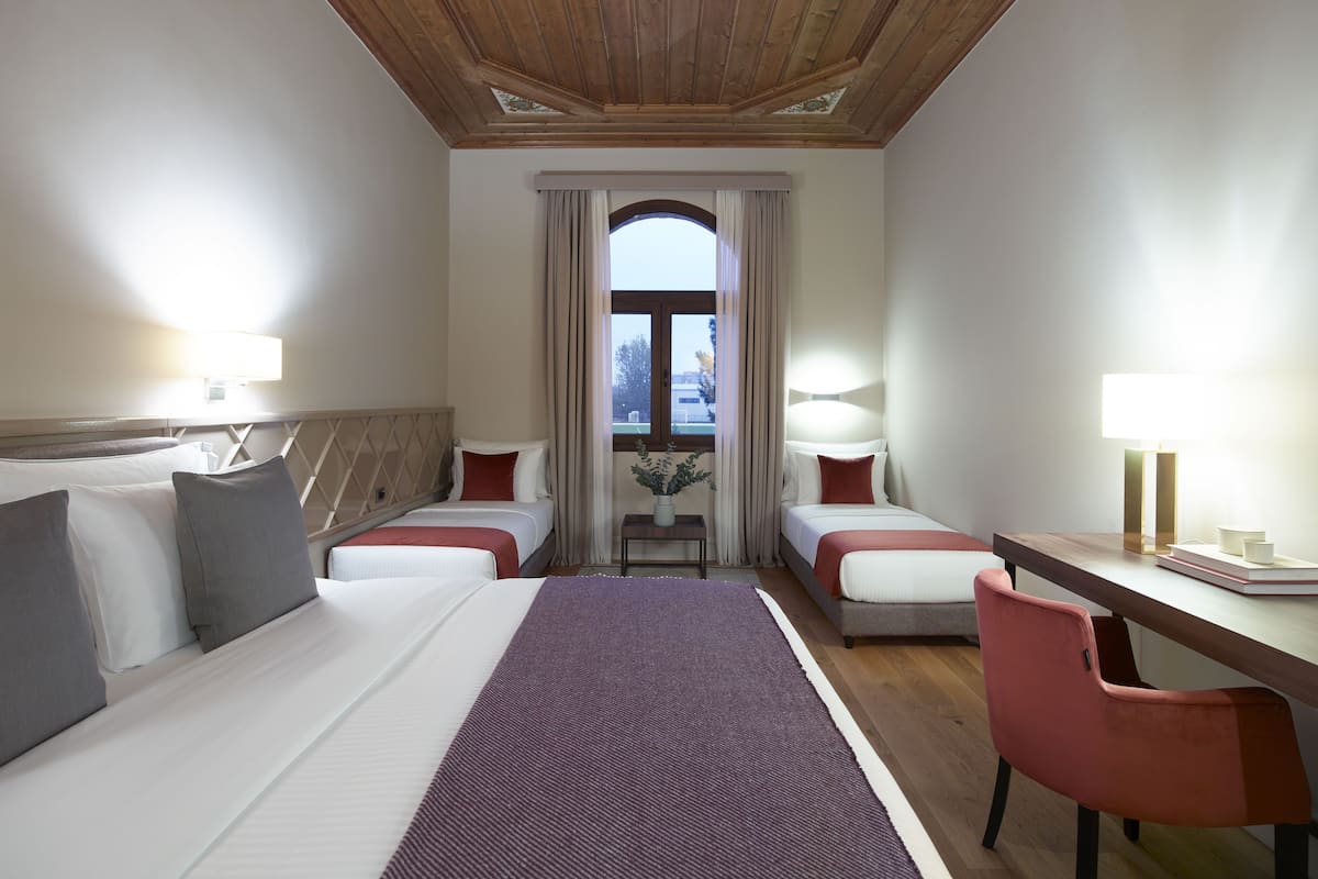 A hotel room with two beds, a desk, and a chair. There is a window with curtains, a plant, and a view of the outside.