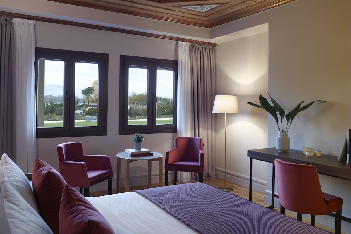 A hotel room with a bed, two chairs, a small table with a plant, a lamp, and a view of the outdoors through large windows.