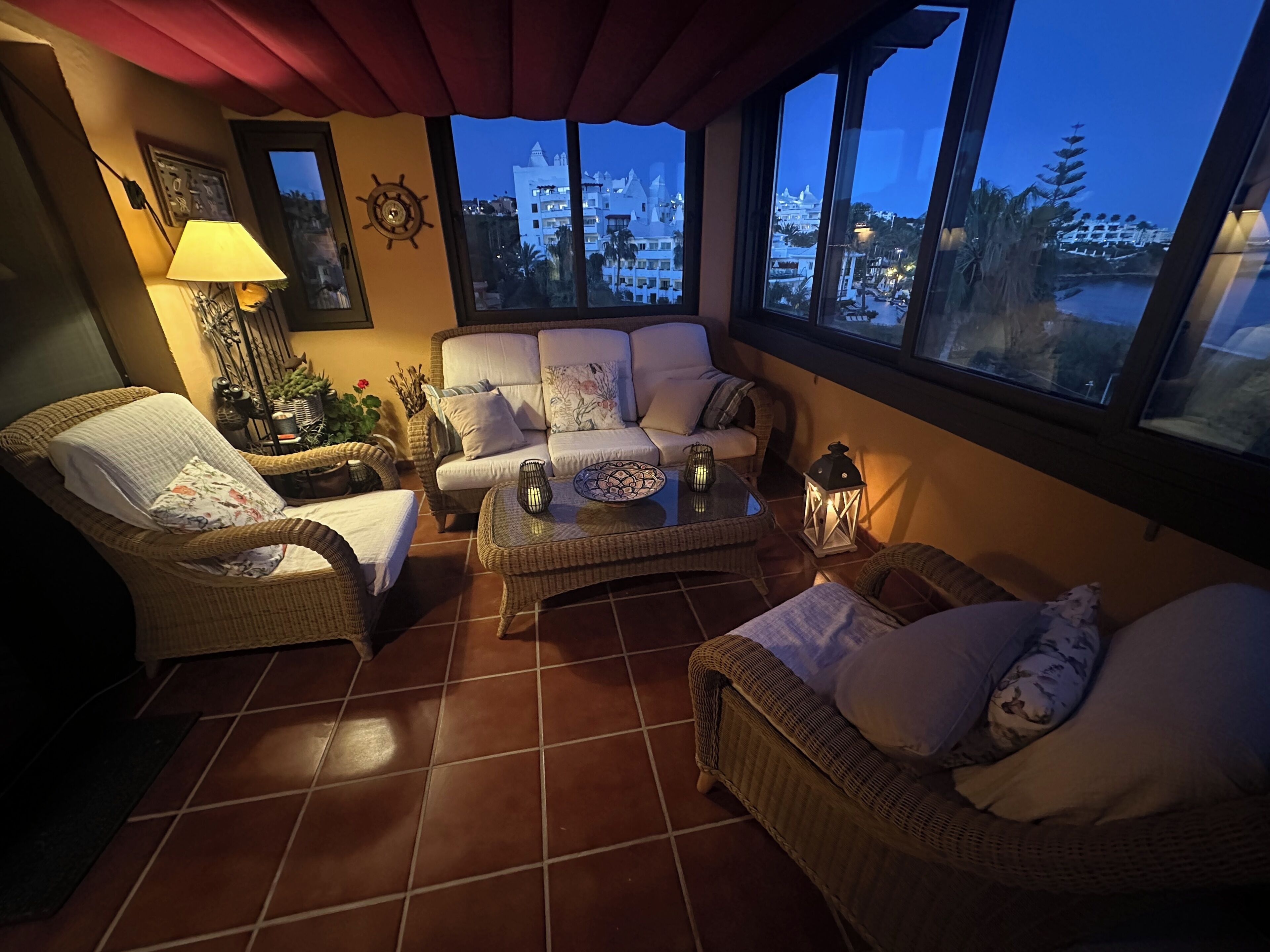 A cozy living room with wicker furniture, a large window with a view of buildings, and a decorative wall clock.