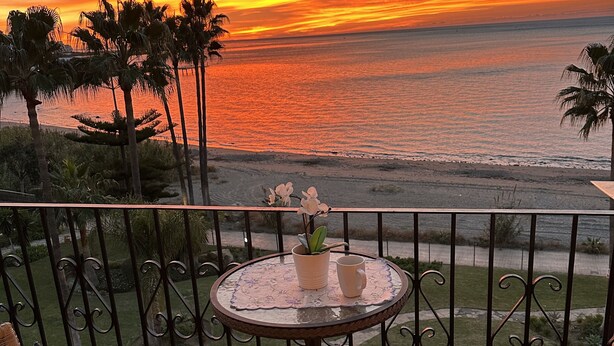 Un balcón con sillas de mimbre y una pequeña mesa, con vista a una playa y al atardecer.