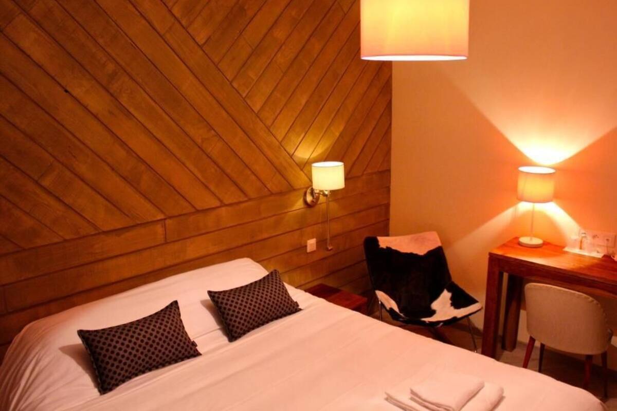 A hotel room with a wooden ceiling, a bed with two pillows, a desk with a lamp, and a chair.