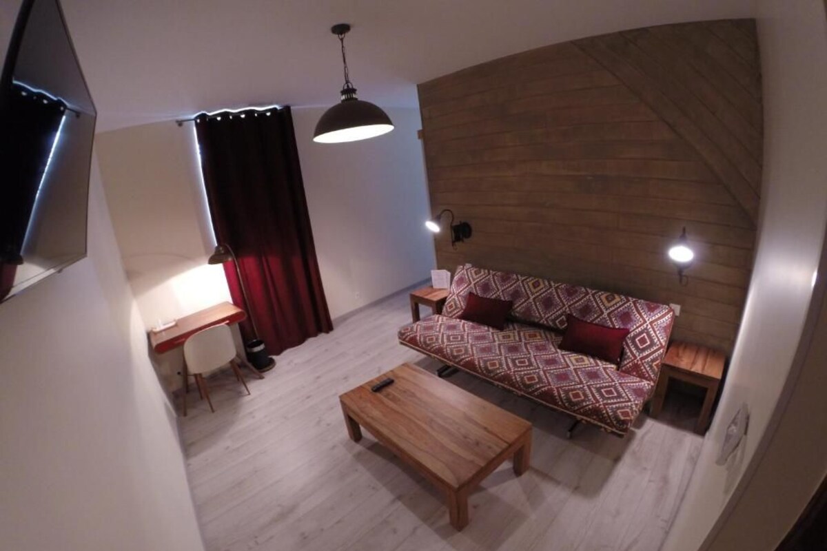 A room with a wooden floor, a sofa with patterned cushions, a wooden coffee table, a desk with a chair, and a wall-mounted TV.