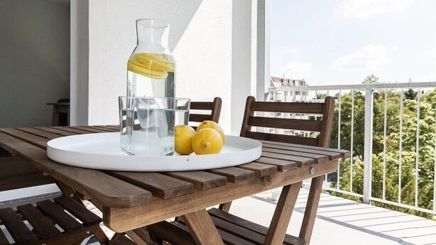 A wooden table with a glass pitcher and lemons on a white tray, set on a balcony with a railing and greenery in the background. - Greystay Apartments Steglitz (Berlin)