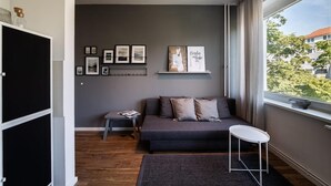 A modern living room with a grey sofa, a small table, and a window with a view of buildings. - Greystay Apartments Steglitz (Berlin)